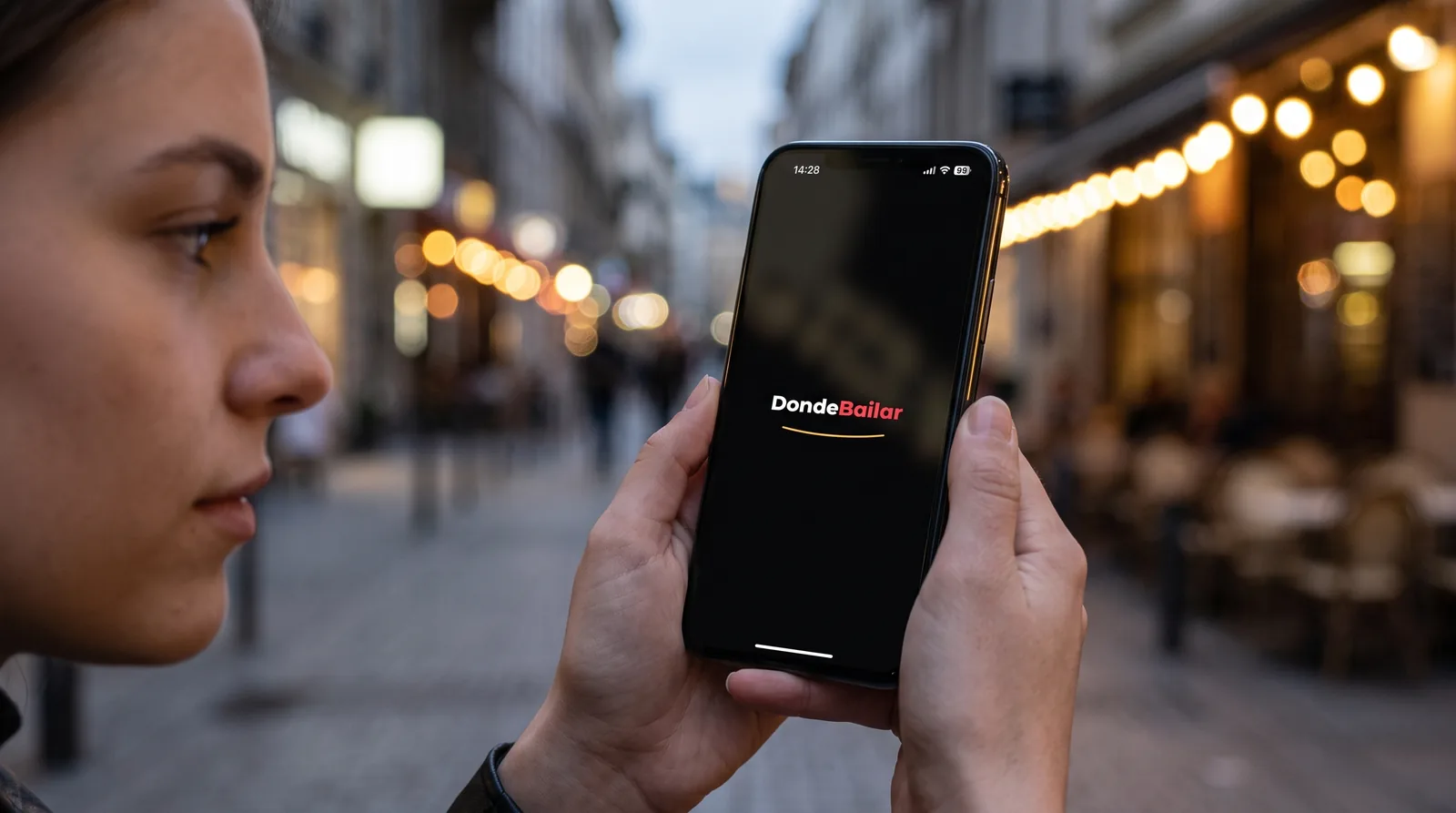 Una persona mira la pantalla de inicio de DondeBailar en su teléfono, en una calle con luces al anochecer